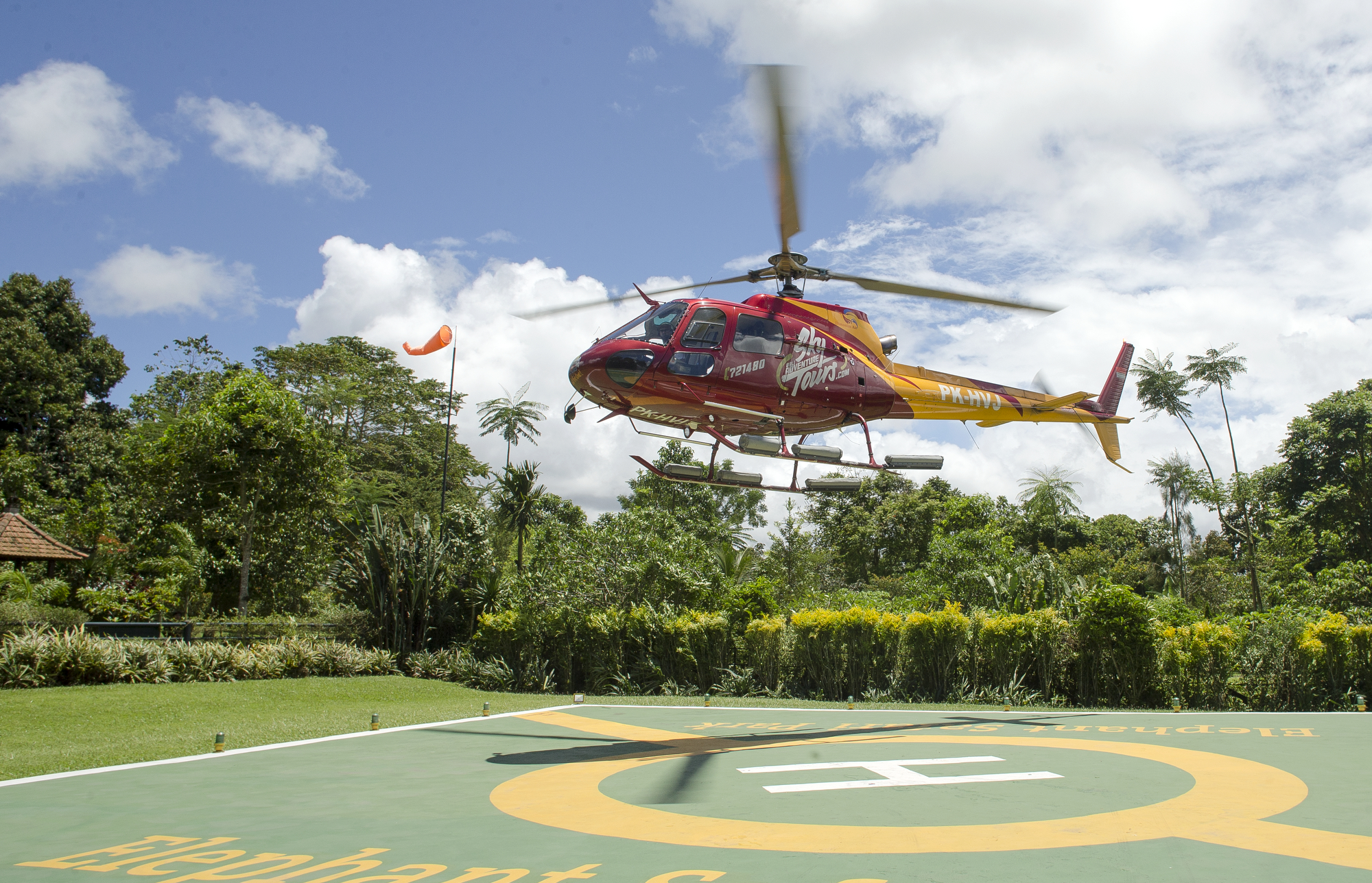 A helicopter glides over a green field, advertising Mason Sky Tours for stunning aerial views of Bali's landscape.