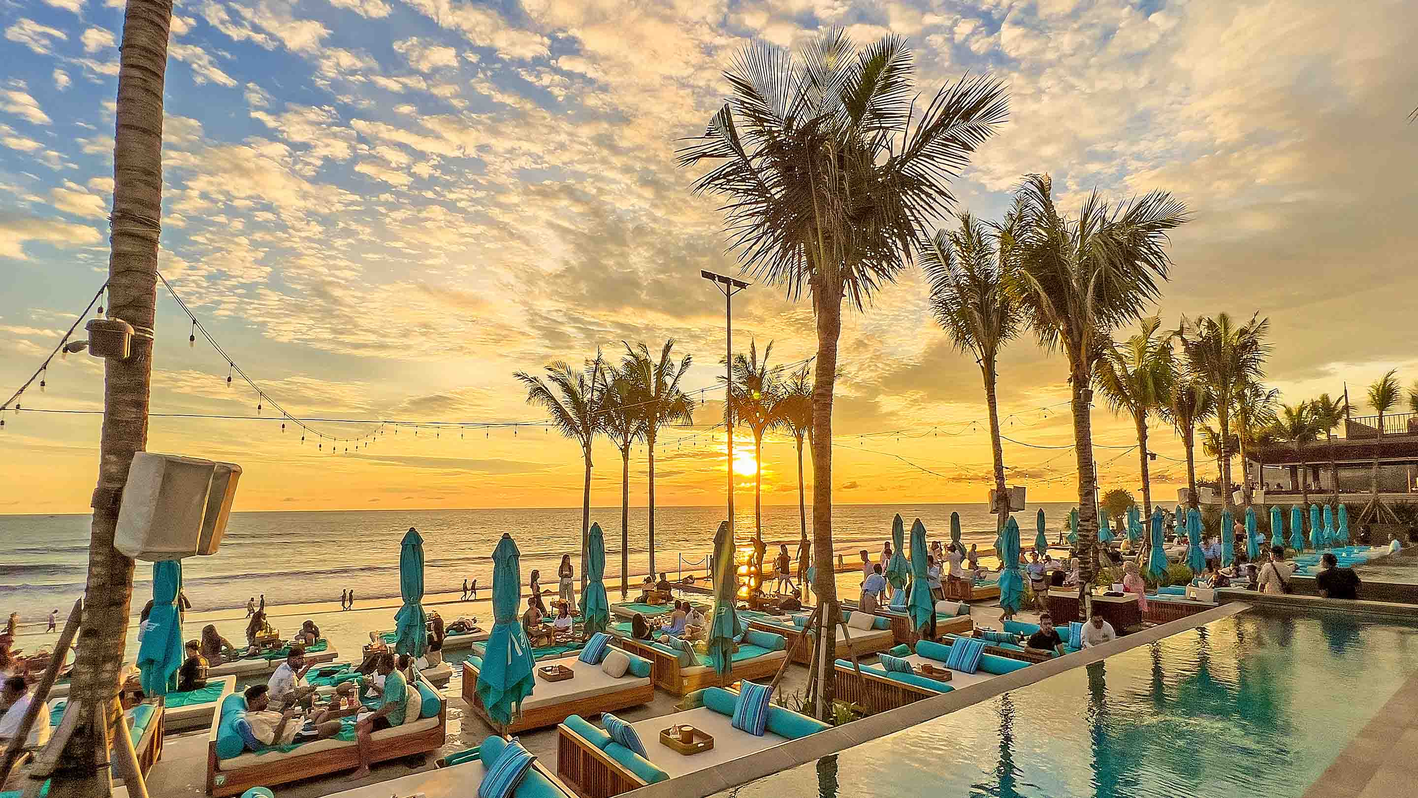 Sunset casts a warm glow over the beach club, with silhouettes of palm trees and lounge chairs lining the shore.